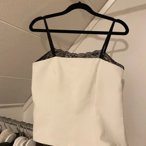 Vit topp med spetsdetaljer - Snygg vit topp med svarta spetsdetaljer upptill. Den har smala axelband och en elegant passform som framhäver figuren. Perfekt för en stilren look🩷