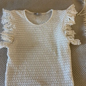 Vit topp med spetsdetaljer från Zara - Säljer en söt vit topp från Zara med spetsdetaljer på ärmarna. Toppen har en texturerad yta och är ärmlös med volangdetaljer. Perfekt för en somrig look! Mycket bra skick 