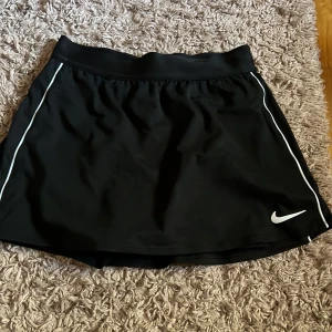 Svart sportkjol från Nike - Snygg svart sportkjol från Nike med Dri-FIT-teknologi för optimal komfort. Kjolen har vita detaljer längs sidorna och en elastisk midja för perfekt passform. Med cykelbyxor inuti. Perfekt för aktiva dagar! Pris kan diskuteras vid snabb affär💞