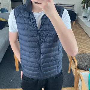 Mörkblå dunväst från Uniqlo - Säljer en stilren mörkblå dunväst från Uniqlo. Västen är ultralätt och har en dragkedja framtill. Perfekt för lager-på-lager under kyliga dagar. Den är designad för att hålla dig varm utan att kännas tung. Passar bra till både casual och mer uppklädda tillfällen.