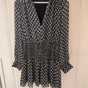 Mönstrad klänning från Zara - Snygg mönstrad klänning från Zara med smockdetaljer i midjan. Klänningen har långa ärmar och ett diskret mönster i svart och vitt. Perfekt för en stilren look.