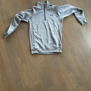 Grå Under Armour - Säljer en stilren grå tröja från Under Armour med halv dragkedja och hög krage. Perfekt för träning eller vardagsbruk. Tröjan har långa ärmar och en diskret logga på bröstet.