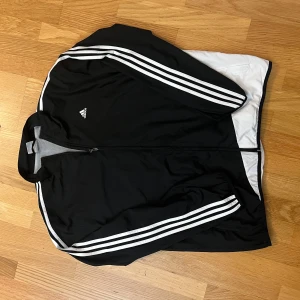 Vintage Adidas Windbreaker Jacka – Svart/Vit (00-tal) - Stilren och klassisk Adidas windbreaker i svart och vitt med ikoniska 3-stripes på ärmarna. Perfekt för en sportig eller retro streetwear-look! Lätt och bekvämt material som funkar året runt. Jackan har dragkedja framtill, två fickor och en skön passform. Adidas-logga på bröstet.  ✔️ Storlek: XL ✔️ Skick: Bra vintage-skick, inga defekter eller fläckar ✔️ Passar perfekt för vår, höst och lager på vintern  ⚡ Snabb leverans & skickas inom 24 timmar! 📩 Fråga gärna om du vill ha fler bilder eller info!