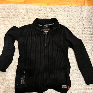 Svart tröja från Sail Racing - Säljer tröja från Sail Racing med dragkedja. Tröjan har långa ärmar och en praktisk ficka med knappar på framsidan. Perfekt för kyligare dagar!