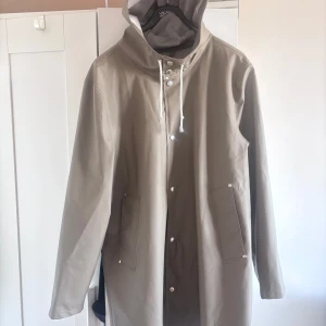 Beige regnjacka från Stutterheim - Säljer en stilren beige regnjacka från Stutterheim. Jackan har en klassisk design med huva och dragsko, samt knappar framtill. Perfekt för regniga dagar och håller dig torr med stil. 🧥
