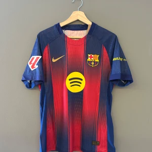 FC Barcelona fotbollströja 25/26 - Snygg FC Barcelona fotbollströja från Nike med klubbens emblem och sponsorloggor. Tröjan har ett klassiskt randigt mönster i rött och blått med gula detaljer. Perfekt för fans som vill visa sitt stöd. En replika helt ny med tagg i storlek M