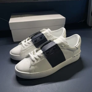 Valentino Garavani sneakers i vitt och svart - Snygga Valentino Garavani sneakers i vitt med en bred svart rem över snörningen. Skorna har en stilren design med subtila detaljer och är tillverkade i skinn. Perfekta för en trendig look. Skriv om ni undrar något box med följer!