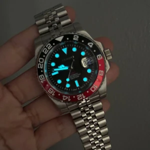 Seiko GMT Pepsi Mod - Inspirerad av den ikoniska GMT-designen och driven av Seiko NH34 automatiska urverk, presenterar vi Seiko Mod GMT—en 40 mm exklusiv klocka som förenar excellens med tillgänglighet. Specifikationer: Diameter 40 mm, tjocklek 12 mm, safirglas, Luminova, 316L rostfritt stål, skruvad krona, keramisk bezel, justerbar handledsstorlek 14,5–22 cm, 20 mm bandbredd, säkerhetsspänne, transparent boettlock, 3 ATM vattentålighet, fullt fungerande GMT-funktion.