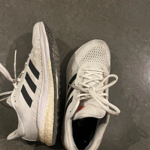 Vita Adidas sneakers med svarta ränder - Snygga vita Adidas sneakers med klassiska svarta ränder. Skorna har snörning och en bekväm sula som ger bra stöd. Perfekta för både träning och vardag.