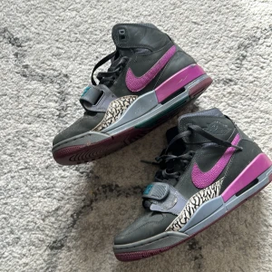 Nike 312 Jordan legacy med lila och turkosa detaljer - Snygga Nike sneakers i svart med lila och turkosa detaljer. Skorna har en hög design med snörning och kardborreband för extra stöd. De har även ett unikt mönster på sidan och en rund tå. Perfekta för en stilren look. Storlek 40, måttligt använda och inga större defekter
