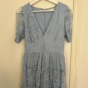 Ljusblå klänning med spetsdetaljer från NLY Trend - Säljer en vacker ljusblå klänning från NLY Trend med spetsdetaljer och blommönster. Klänningen har en v-ringad design och korta ärmar med elastiska avslut. Perfekt för sommaren och knappt använd 
