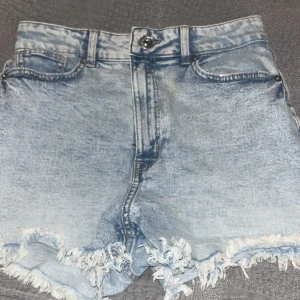 Blå jeansshorts med fransar - Snygga blå jeansshorts med fransiga kanter och hög midja. Perfekta för sommardagar och ger en avslappnad look. De har klassisk femficksdesign och en dragkedja framtill.