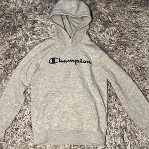 Grå hoodie från Champion - En ljusgrå hoodie från Champion med deras  logga på bröstet. Perfekt för en avslappnad dag eller annat, storlek L som ska passa 11/12 åringar som är mellan 150 till 155cm. Minimalt använd. Hör gärna av er om frågor eller annat 😇✨