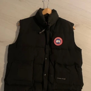 Svart dunväst från Canada Goose - Säljer en svart dunväst från Canada Goose med deras ikoniska märke på bröstet. Västen har en dragkedja och knappar framtill samt praktiska fickor. Perfekt för kyliga dagar. !OBS! Västen är är liten i storlek, så rekommenderar starkt för XS/S! Två knappar är även sönder men inget som syns!