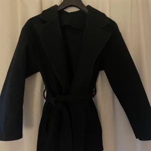 Svart kappa med bälte - Snygg svart kappa med bälte som ger en elegant look. Perfekt för kyligare dagar och passar till många olika outfits. Kappan har långa ärmar och en klassisk krage.