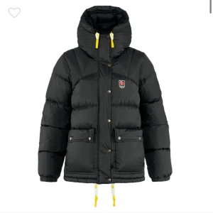 Svart dunjacka från Fjällräven - Säljer en svart dunjacka från Fjällräven med gula detaljer. Jackan har en hög krage och dragkedja med knappar framtill. Den är utrustad med praktiska fickor och justerbara snören vid huvan och nederkanten. Skriv för fler bilder så löser jag❤️