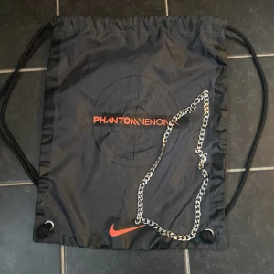 Svart gympåse från Nike - Snygg svart gympåse från Nike med texten 'Phantom Venom' i orange. Påsen har en praktisk dragsko och en kedja som detalj. Perfekt för att bära med sig träningskläder eller andra småsaker.