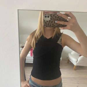 Svart ärmlös topp med öppen rygg - Snygg svart ärmlös topp med en unik design och öppen rygg. Perfekt för en stilren look. Toppen har en omlott-detalj framtill som ger en extra touch.