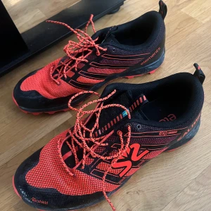 Svart och röd träningssko från Salming - Säljer ett par svarta och röda träningsskor från Salming med snörning och mönstrad ovandel. Skorna har en sportig design med Salming-logga och är perfekta för löpning eller annan träning.