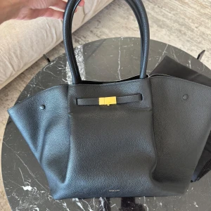 Svart handväska från DeMellier - New York har en modern men tidlös siluett, skuren av hållbara material med vår distinkta bältesdetalj. De långa topphandtagen svänger lätt över axeln eller kan öglas över armkroken. Essentials är säkert säkrade i en rymlig bomullsfodrad insida med flera fickor, stängda med en central magnet och två tryckknappar på sidorna. New York är den perfekta dag-nattväskan inspirerad av vår kärlek till staden som aldrig sover.  Inredning: Huvudfack med magnetisk stängning lämplig för A4-filer eller bärbar 