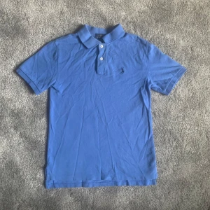 Blå pikétröja från Ralph Lauren - Säljer en klassisk blå pikétröja från Ralph Lauren med broderad logga på bröstet. Tröjan har korta ärmar och knappar vid kragen. Perfekt för en stilren look!