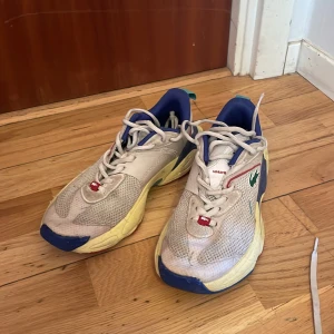 Färgglada sneakers från Lacoste - Säljer ett par unika sneakers från Lacoste med en färgglad design. Skorna har en vit bas med inslag av blått, rött och grönt. De har snörning och en rund tå, samt en mönstrad sula i flera färger. Perfekta för dig som vill sticka ut!