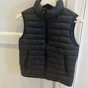 Svart dunväst med dragkedja - Snygg svart dunväst med dragkedja och hög krage. Perfekt för kyliga dagar och enkel att matcha med olika outfits. Två praktiska fickor med dragkedja framtill.