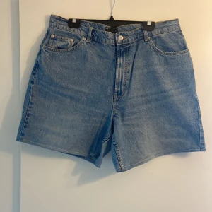 Blå jeansshorts från ASOS DESIGN - Klassiska blå jeansshorts från ASOS DESIGN med fem fickor och hög midja. Perfekta för sommaren med en avslappnad passform och tidlös denimlook. Stängs med dragkedja och knapp framtill. Obs! Har sytt om dessa från jeans så dem är inte helt jämna (som fabrikssydda shorts) och har en söm i vit tråd.