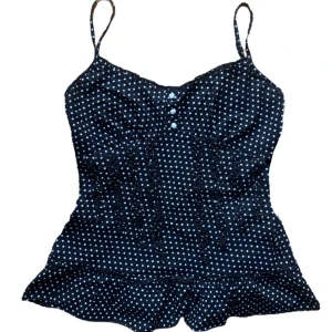 Svart prickig linnetopp med knappar - Supersöt svart linnetopp med vita prickar och smala axelband. Toppen har dekorativa knappar framtill och volangkant nertill. Tillverkad i 100% bomull vilket gör den sval och skön. 