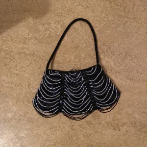 Svart handväska med pärldetaljer - Säljer en svart handväska med pärlbroderade vågmönster på framsidan. Väskan har ett handtag och stängs med dragkedja. Perfekt för att addera lite extra till din outfit!