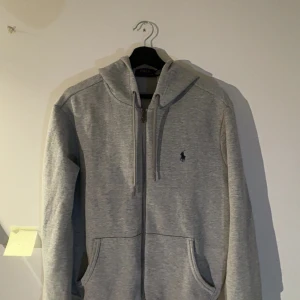 Grå hoodie från Polo Ralph Lauren - Säljer en klassisk grå hoodie från Polo Ralph Lauren med dragkedja och huva. Tröjan har två fickor framtill och en liten broderad logga på bröstet. Perfekt för en avslappnad stil. Som ny