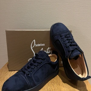 Mörkblå mocka sneakers från Christian Louboutin - Säljer ett par mörkblå sneakers i mocka från Christian Louboutin med klassisk röd sula. Skorna har rund tå, platt sula och snörning. Perfekta för dig som vill ha en stilren och lyxig look.
