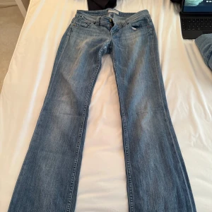 Blå bootcut low waist jeans - Säljer ett par klassiska blå bootcut jeans med slitna detaljer och fransar vid bensluten. Jeansen har normal passform och stängs med dragkedja och knapp. Perfekta för dig som gillar en avslappnad och vintage-inspirerad stil.