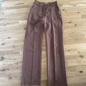 Bruna kostymbyxor från Madlady - Säljer ett par bruna kostymbyxor från Madlady med raka ben och pressveck. Byxorna har fickor framtill och en klassisk passform som passar till många olika stilar.