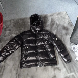 Svart dunjacka med huva från Moncler - Säljer en svart glansig dunjacka från Moncler med huva och dragkedja. Jackan har klassisk pufferdesign, två fickor framtill och Moncler-logga på ärmen. Perfekt för kyliga dagar och riktigt snygg streetstil.