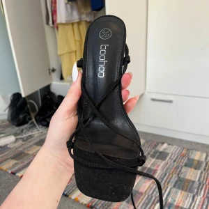 Svarta klackskor med snörning från Boohoo - Säljer ett par svarta klackskor från Boohoo med smala remmar och snörning som går att knyta runt vristen. Skorna har fyrkantig tå och hög klack. Superfina men passa tyvärr inte mig💗