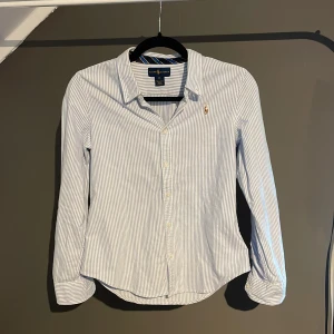 Blåvit randig skjorta från Ralph Lauren - Klassisk skjorta från Ralph Lauren i blåvitt randigt mönster. Skjortan har långa ärmar, knappar framtill och en broderad logga på bröstet. Perfekt för en stilren och tidlös look.