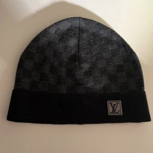  Lv mössa - Snygg svart mössa från Louis Vuitton med diskret rutigt mönster och uppvikt kant. Märkets logotyp sitter framtill på kanten. Perfekt accessoar för dig som gillar stilrena detaljer.