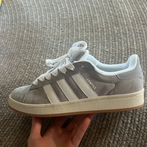 Adidas Campus ljusgrå sneakers - Säljer ett par Adidas Campus sneakers i ljusgrå mocka med vita detaljer och klassiska tre ränder på sidan. Skorna har vit sula, snörning och Adidas-logga på hälen. Perfekta för en stilren och avslappnad look. Skorna är inte använde utan bara köpt av en kompis men säljs eftersom de var lite små. Lite små i storleken, skulle passa 43 eller 42,5