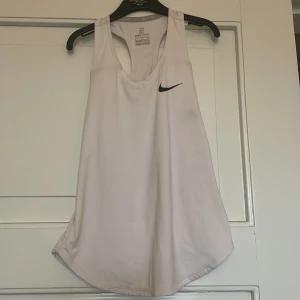 Vit Dri-FIT sporttop från Nike - Säljer en vit sportig linnetopp från Nike med Dri-FIT-teknologi. Toppen har brottarrygg och ett svart Nike Swoosh-tryck på bröstet. Perfekt för träning och aktiva dagar.
