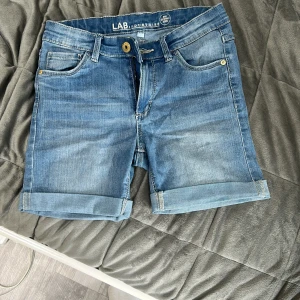 Blå jeansshorts från LAB Industries - Snygga blå jeansshorts från LAB Industries med klassisk femficksdesign och uppvikta benslut. Shortsen har gylf med dragkedja och knapp samt bälteshällor. Perfekta för varma dagar!