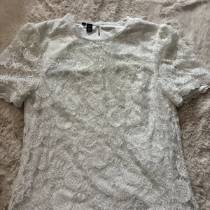 Vit spetsblus  - Säljer en söt vit spetsblus med korta ärmar och rund halsringning. Blusen har fina spetsdetaljer och ett diskret mönster över hela tyget. Perfekt att styla med jeans eller kjol för en fräsch look.💕