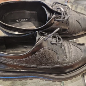 Svarta läderskor från Tiger of Sweden - Säljer ett par svarta läderskor från Tiger of Sweden med snörning och rund tå. Skorna har en platt sula med blå detalj och klassisk design, perfekta för en stilren look.