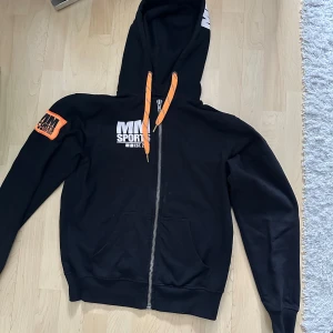 MM Sports svart hoodie - Svart hoodie från MM Sports med vit logga på bröstet och huvan. Orange detaljer på dragsko och patch på ärmen. Dragkedja framtill och två fickor. Perfekt för träning eller chill.