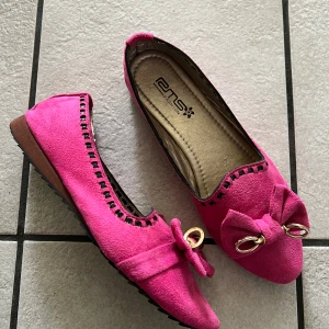 Rosa ballerinaskor med rosett och gulddetaljer - Säljer ett par knallrosa ballerinaskor från EMS Design i mocka-look. Skorna har en stor rosett framtill med guldfärgade ringar som detalj och svarta dekorativa sömmar längs kanten. Platt sula och rund tå för en bekväm stil.