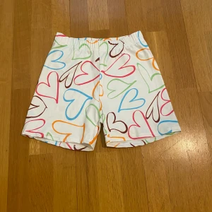 Shorts - Säljer ett par vita shorts med färgglada hjärtan i olika nyanser. De har en elastisk midja och är tillverkade i mjuk och stretchig polyesterblandning. Perfekta för sommarens alla äventyr!