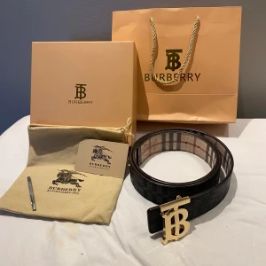 Burberry bälte med rutigt mönster och guldfärgat spänne - Säljer ett Burberry bälte med klassiskt rutigt mönster i beige, svart och rött. Bältet har ett stort guldfärgat 'TB'-spänne och är tillverkat i skinn. Perfekt accessoar för att lyfta din outfit.