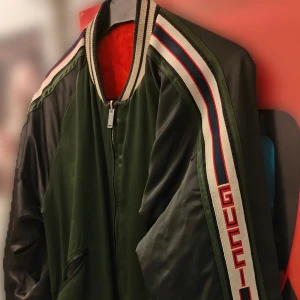 Grön/Orange jacka från Gucci - Två jackor i en! Ena sidan är mörkgrön och svart med röda och vita ränder längs ärmarna och logotypen på sidan. Andra sidan är orange. Jackan har dragkedja och ribbade muddar vid krage och ärmslut. Perfekt för dig som vill sticka ut med en exklusiv streetstyle-look.