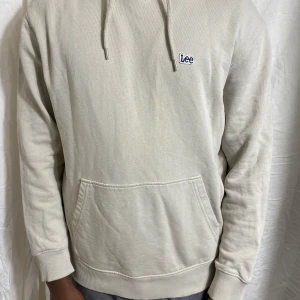 Beige hoodie från Lee - Säljer en stilren beige hoodie från Lee med broderad logga på bröstet. Tröjan har huva med dragsko och en stor magficka. Perfekt för en avslappnad look.