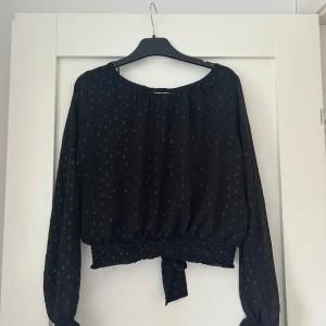 En svart blus - Säljer en svart blus med silvriga prickar och långa ärmar. Blusen har resår och knytning i midjan samt lätt transparent tyg. Perfekt för dig som gillar trendiga detaljer och en feminin look. Storleken är 158 cm/12-13 år men den passar för Xs samt en mindre S. Den är enbart använd under ett tillfälle.